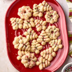 A red platter holds spritz cookies in festive shapes, topped with red, green, and white sprinkles. More cookies, sprinkles, and a metal cookie press are nearby on a light-colored surface.