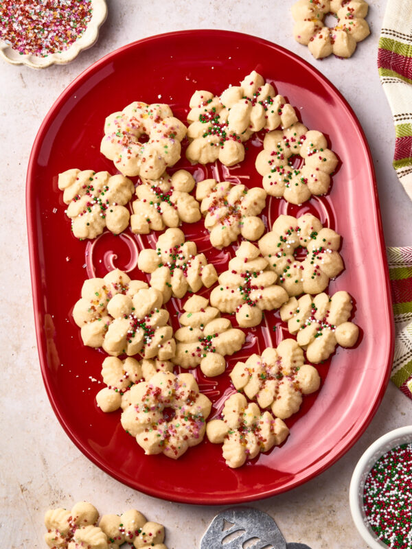 A red platter holds spritz cookies in festive shapes, topped with red, green, and white sprinkles. More cookies, sprinkles, and a metal cookie press are nearby on a light-colored surface.