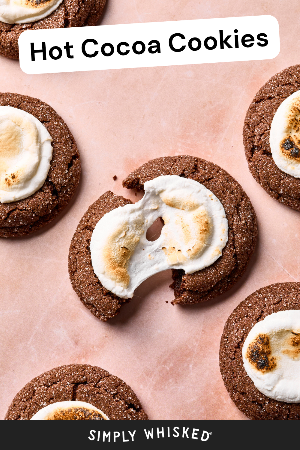 Five hot cocoa cookies, each topped with toasted marshmallow, are arranged on a light pink surface. One cookie in the center is broken in half, revealing its soft interior. Text reads Hot Cocoa Cookies and Simply Whisked.