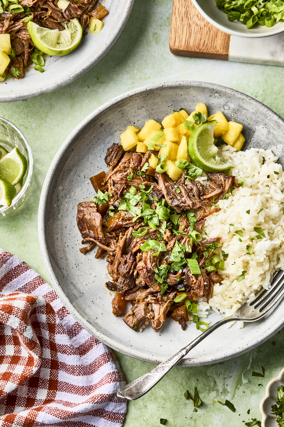 A plate with shredded beef, white rice, pineapple chunks, lime wedges, and chopped cilantro, set on a green surface with a fork, a red checked napkin, and additional plates and garnishes nearby.