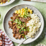 A plate with shredded beef, white rice, pineapple chunks, lime wedges, and chopped herbs, garnished with green onions. A fork rests on the plate. Dishes of green onions and chopped herbs are nearby on a green surface.