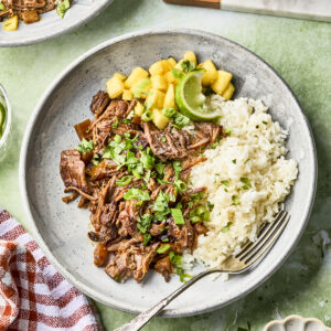 A plate with shredded beef, white rice, pineapple chunks, lime wedges, and chopped herbs, garnished with green onions. A fork rests on the plate. Dishes of green onions and chopped herbs are nearby on a green surface.
