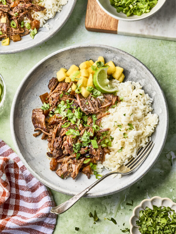 A plate with shredded beef, white rice, pineapple chunks, lime wedges, and chopped herbs, garnished with green onions. A fork rests on the plate. Dishes of green onions and chopped herbs are nearby on a green surface.