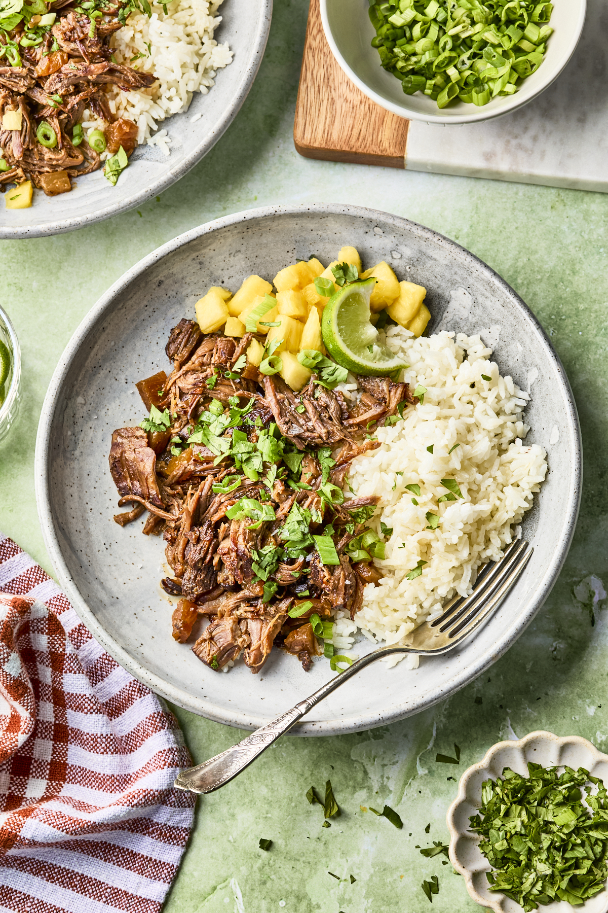 A plate with shredded beef, white rice, pineapple chunks, lime wedges, and chopped herbs, garnished with green onions. A fork rests on the plate. Dishes of green onions and chopped herbs are nearby on a green surface.
