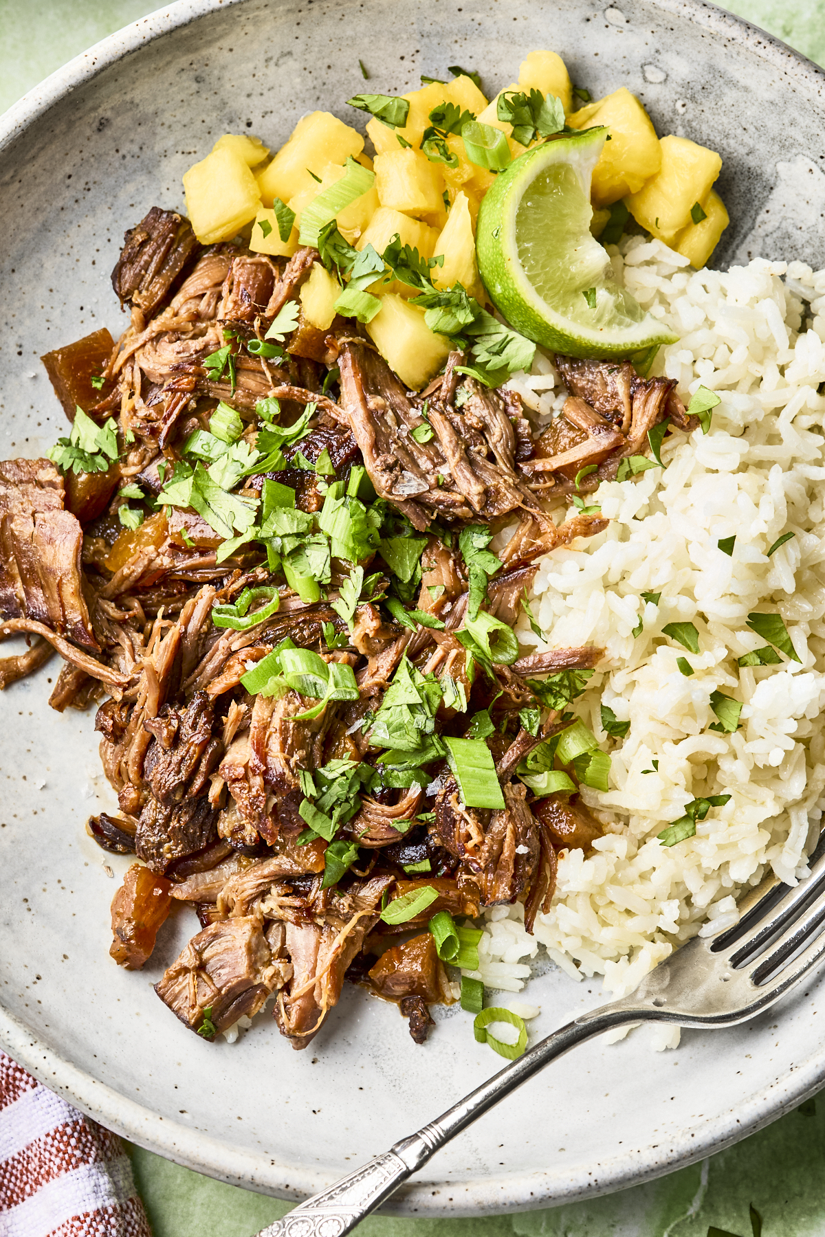 A plate with shredded beef topped with chopped herbs, white rice, pineapple chunks, and a lime wedge, served with a fork on the side.