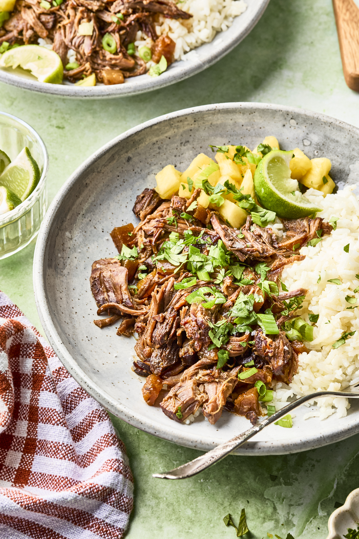 A plate with shredded beef topped with chopped herbs, white rice, pineapple salsa, and lime wedges on a speckled gray dish. A cloth napkin, spoon, and a small bowl of lime wedges are beside the plate.