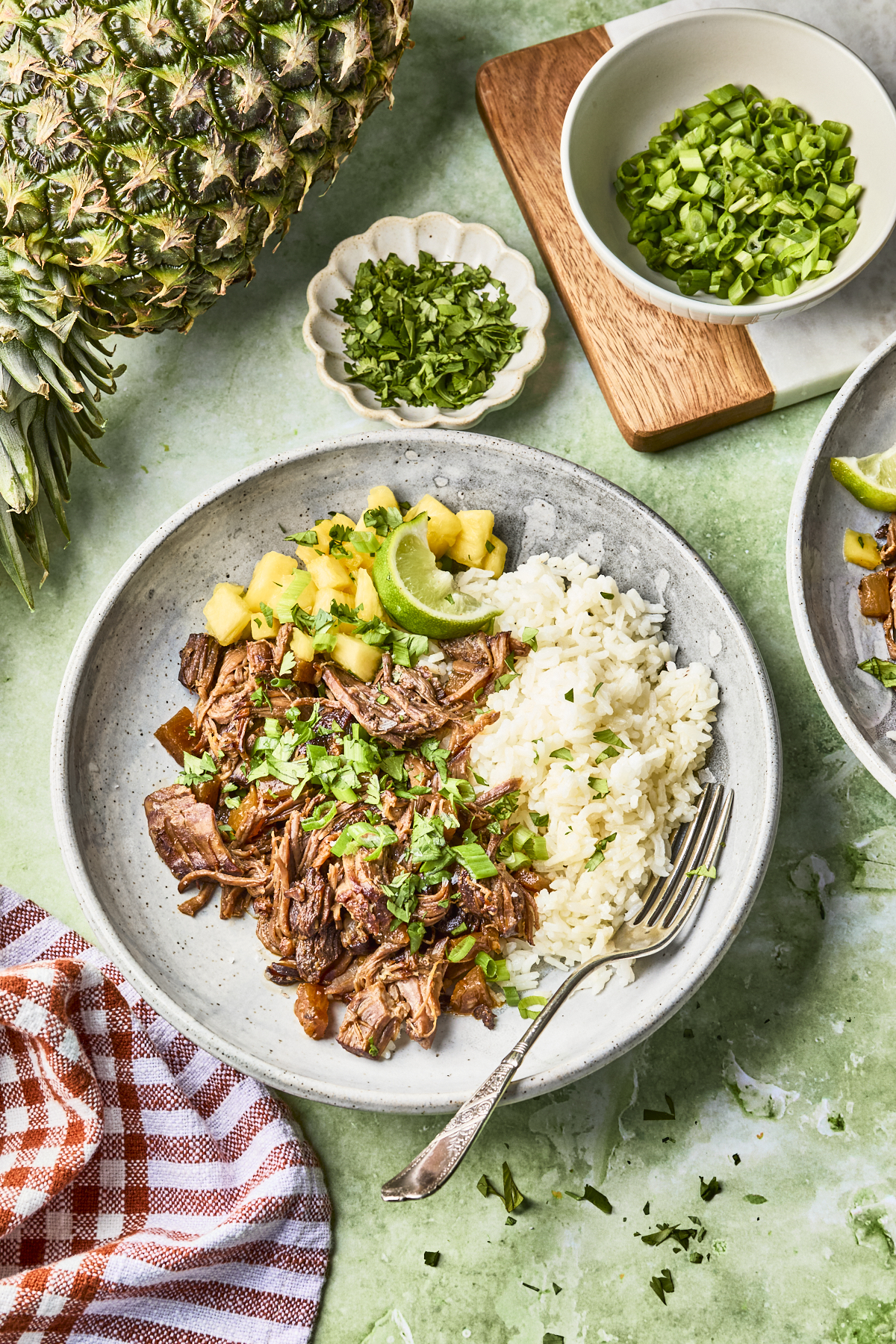 A plate with shredded beef garnished with cilantro, pineapple chunks, lime wedges, and a serving of white rice. Fresh herbs and sliced green onions are in small bowls nearby, alongside a pineapple and a fork on the plate.