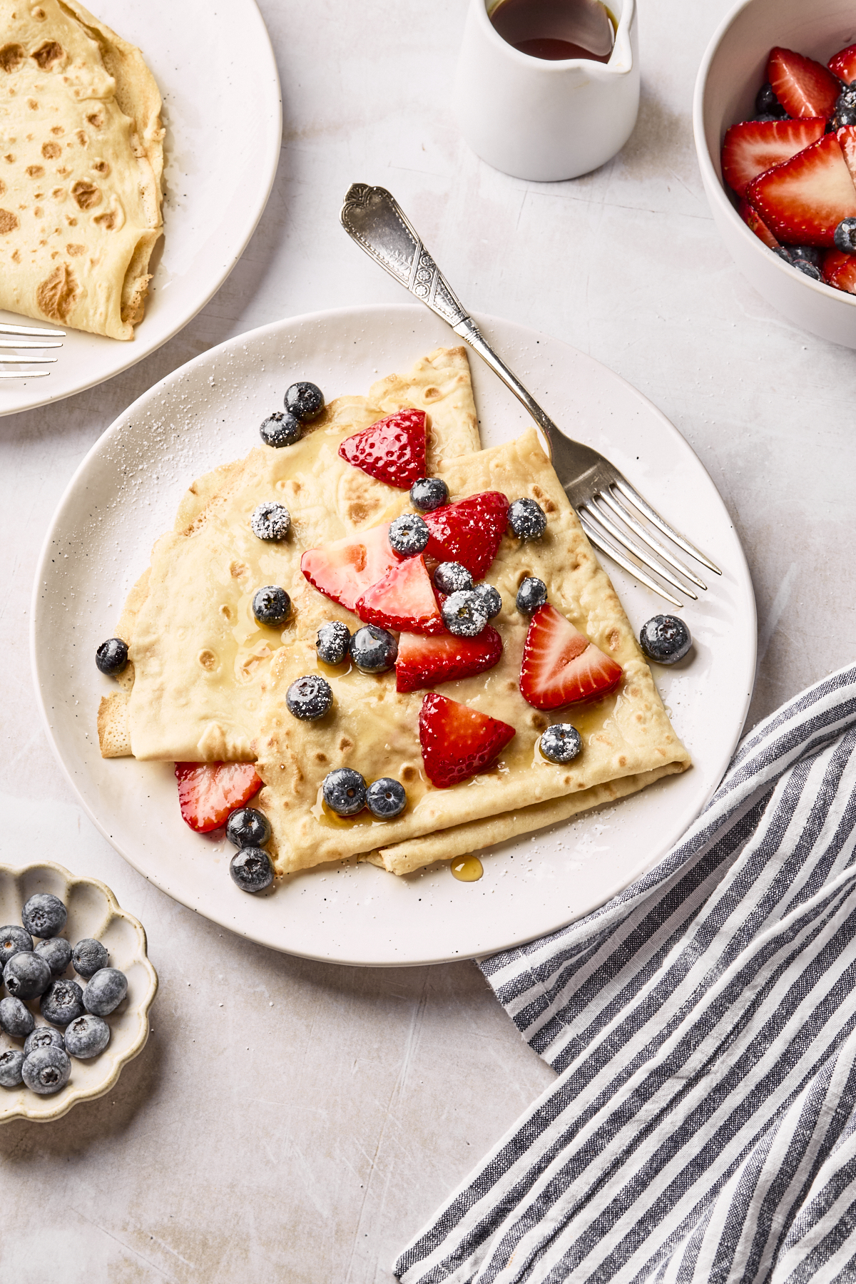 A plate with folded crepes topped with sliced strawberries, blueberries, and syrup. A fork rests on the plate. Surrounding the plate are a bowl of berries, a striped napkin, a pitcher of syrup, and another plate with crepes.