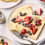 A plate of crepes topped with fresh blueberries, sliced strawberries, and powdered sugar. A fork rests on the plate, with more crepes and berries visible nearby, along with a small pitcher of syrup.