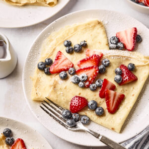 A plate of crepes topped with fresh blueberries, sliced strawberries, and powdered sugar. A fork rests on the plate, with more crepes and berries visible nearby, along with a small pitcher of syrup.