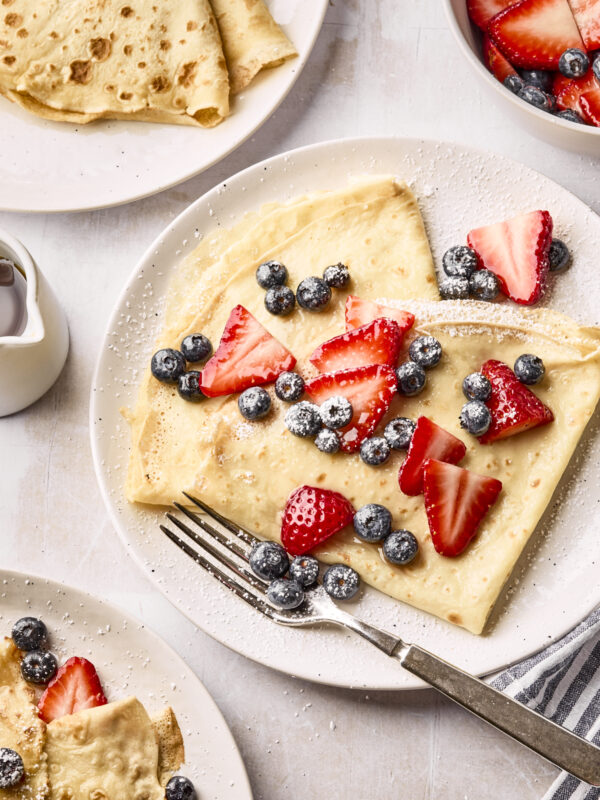 A plate of crepes topped with fresh blueberries, sliced strawberries, and powdered sugar. A fork rests on the plate, with more crepes and berries visible nearby, along with a small pitcher of syrup.