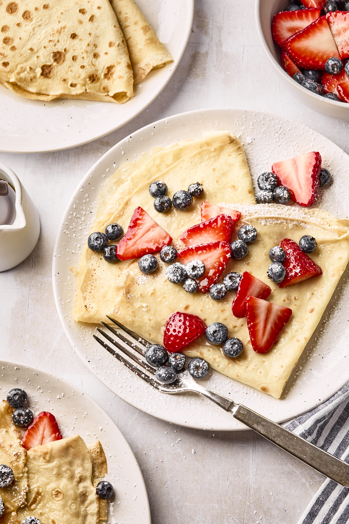 A plate of crepes topped with fresh blueberries, sliced strawberries, and powdered sugar. A fork rests on the plate, with more crepes and berries visible nearby, along with a small pitcher of syrup.