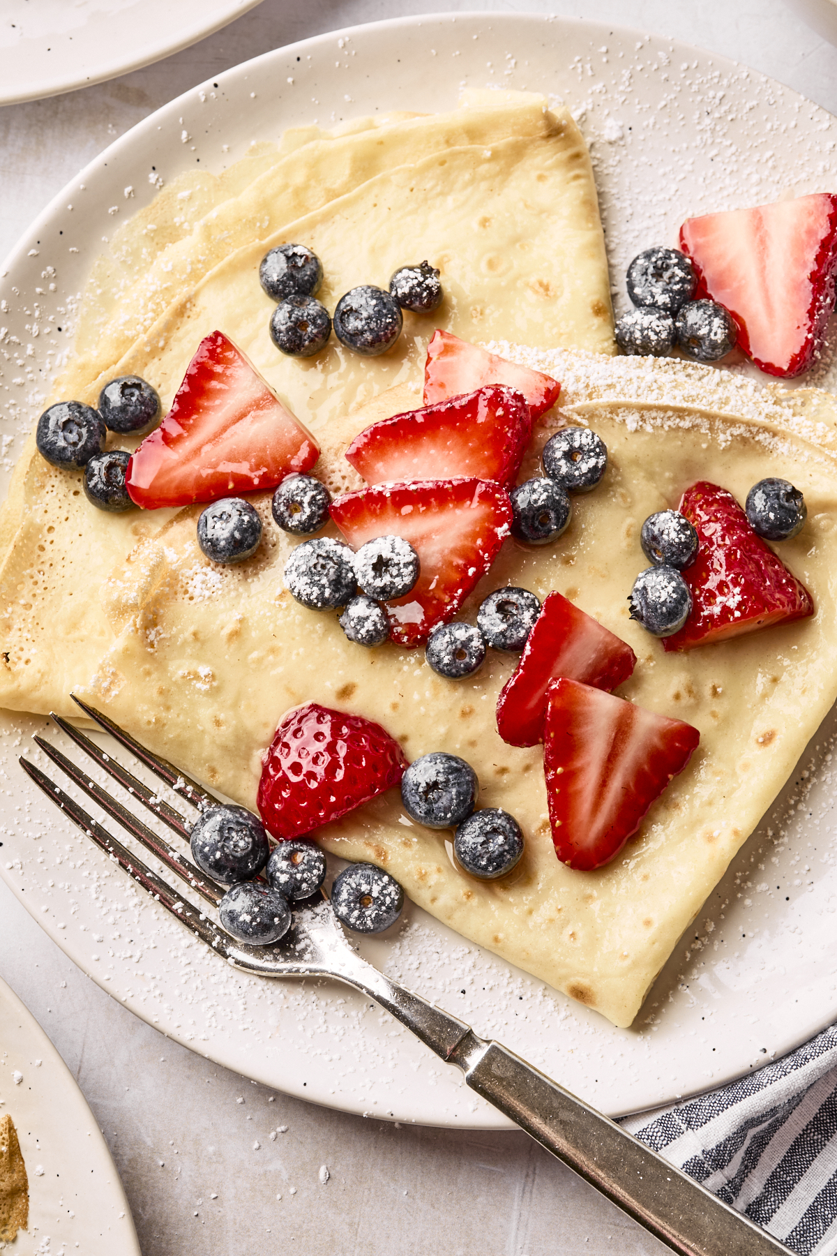 A plate with a folded crepe topped with fresh sliced strawberries, blueberries, and a dusting of powdered sugar. A fork rests on the plate.