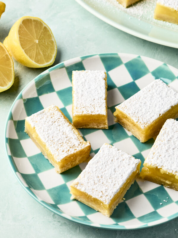 Rectangular lemon bars dusted with powdered sugar are arranged on a green and white checkered plate. Fresh lemon halves sit nearby on a light turquoise surface.