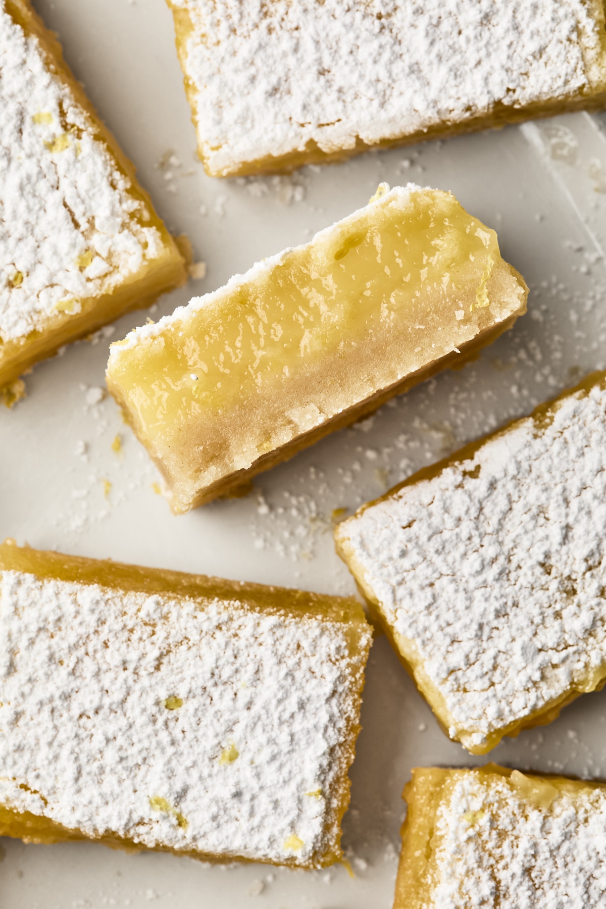 Lemon bars topped with powdered sugar are arranged on a white surface. One bar is turned to show its creamy, yellow filling and buttery crust.