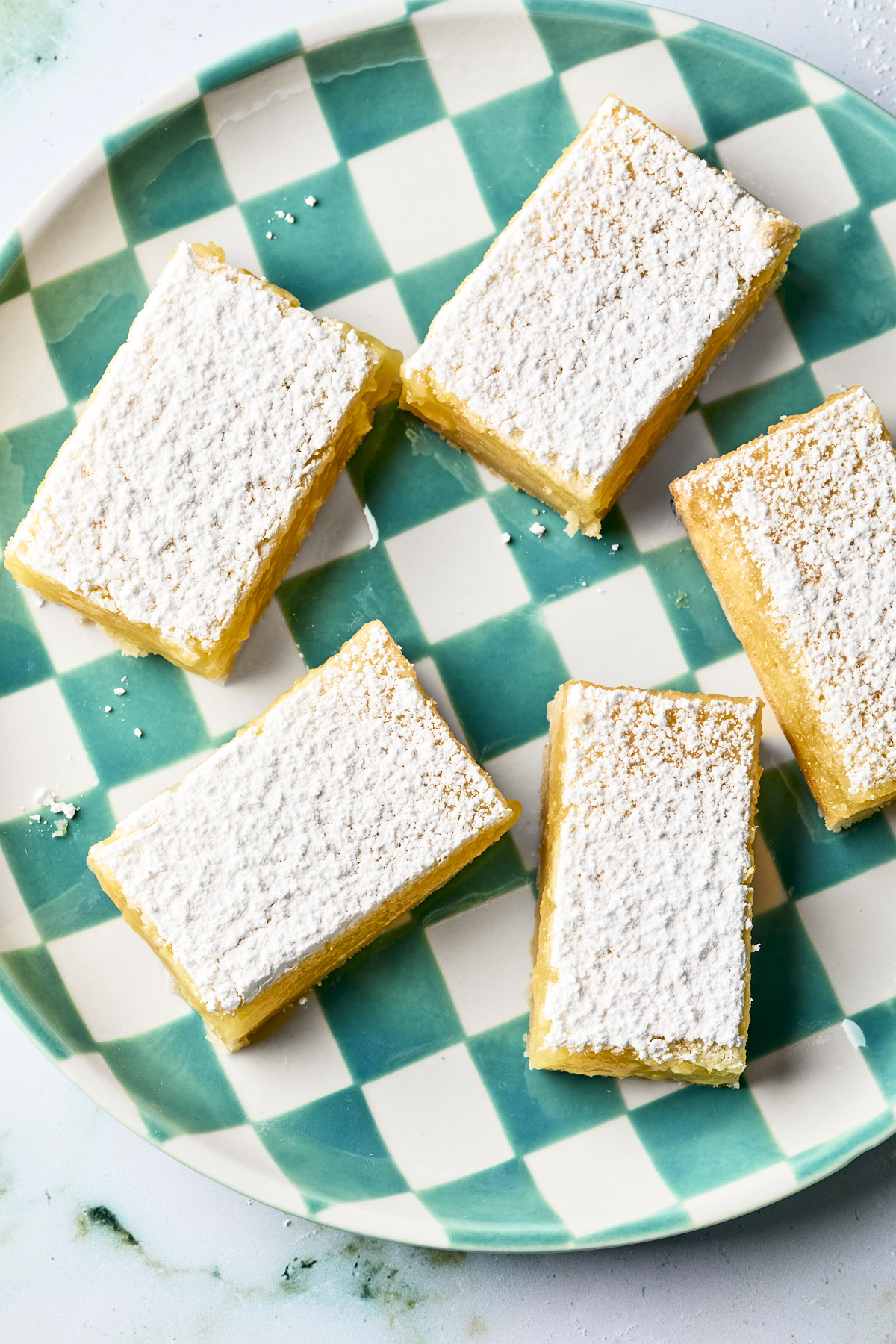 Five lemon bars topped with powdered sugar are arranged on a green and white checkered plate.