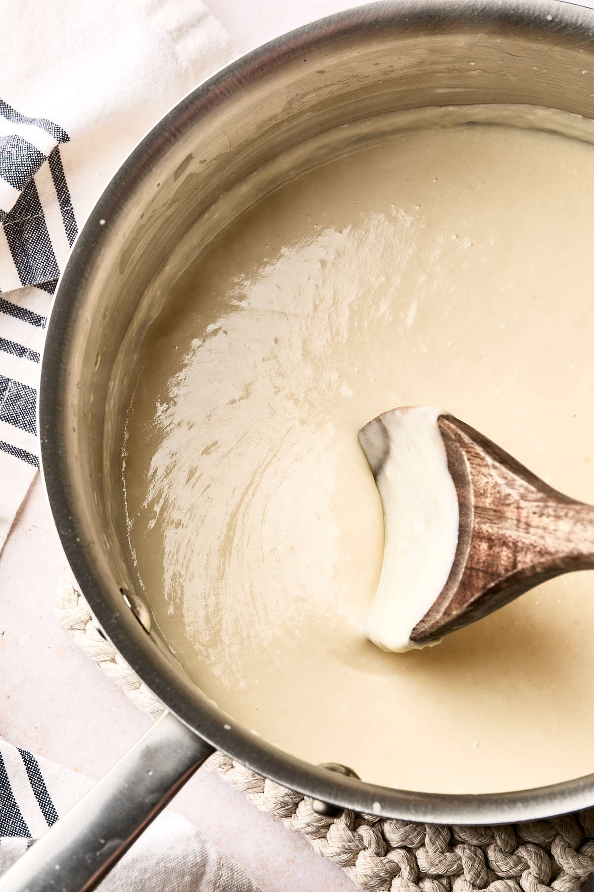 A wooden spoon stirs creamy white sauce in a stainless steel saucepan, with a striped kitchen towel nearby on a light surface.