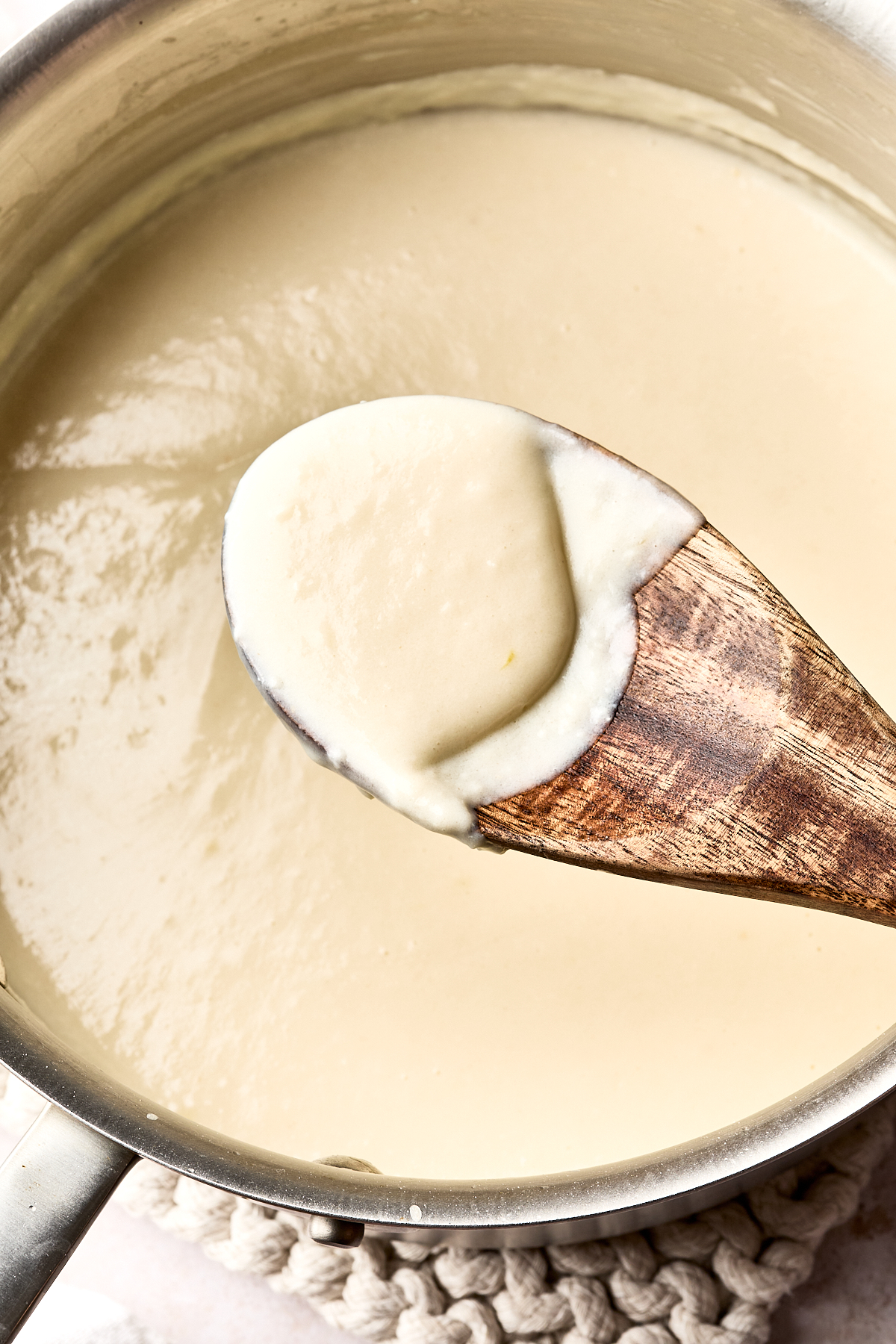 A wooden spoon holds creamy white sauce above a saucepan filled with the same smooth, thick sauce.