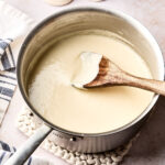 A saucepan filled with creamy white sauce is being stirred with a wooden spoon. The pot sits on a knitted trivet, with salt and pepper shakers and a striped kitchen towel nearby.