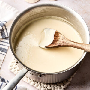 A saucepan filled with creamy white sauce is being stirred with a wooden spoon. The pot sits on a knitted trivet, with salt and pepper shakers and a striped kitchen towel nearby.