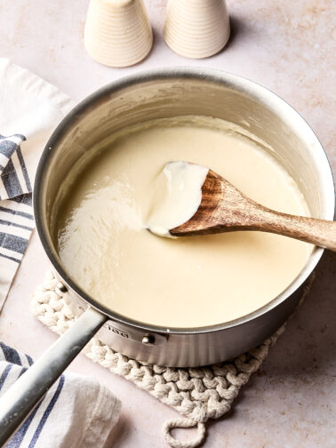 A saucepan filled with creamy white sauce is being stirred with a wooden spoon. The pot sits on a knitted trivet, with salt and pepper shakers and a striped kitchen towel nearby.