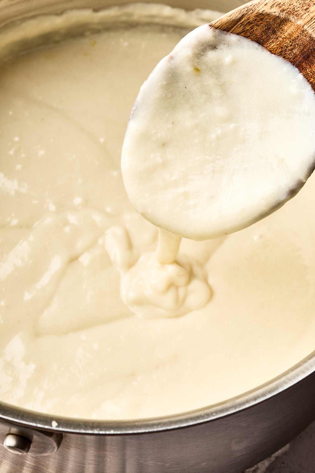 A close-up of creamy white sauce being stirred with a wooden spoon in a metal saucepan. The sauce appears thick and smooth, coating the spoon as it drips back into the pot.