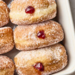 Sugar-coated jelly-filled doughnuts arranged in a white baking dish, each with a spot of red jelly oozing from their centers.