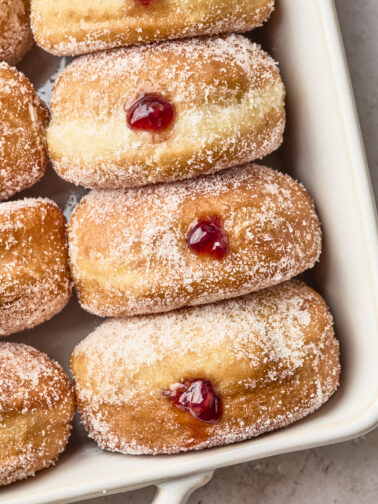 Sugar-coated jelly-filled doughnuts arranged in a white baking dish, each with a spot of red jelly oozing from their centers.