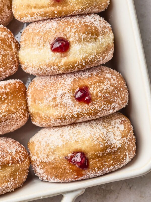 Sugar-coated jelly-filled doughnuts arranged in a white baking dish, each with a spot of red jelly oozing from their centers.