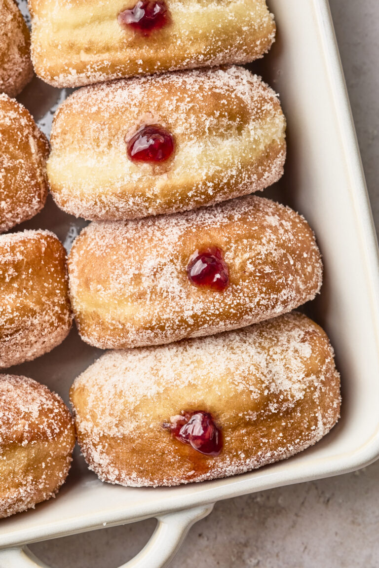 Sugar-coated jelly-filled doughnuts arranged in a white baking dish, each with a spot of red jelly oozing from their centers.