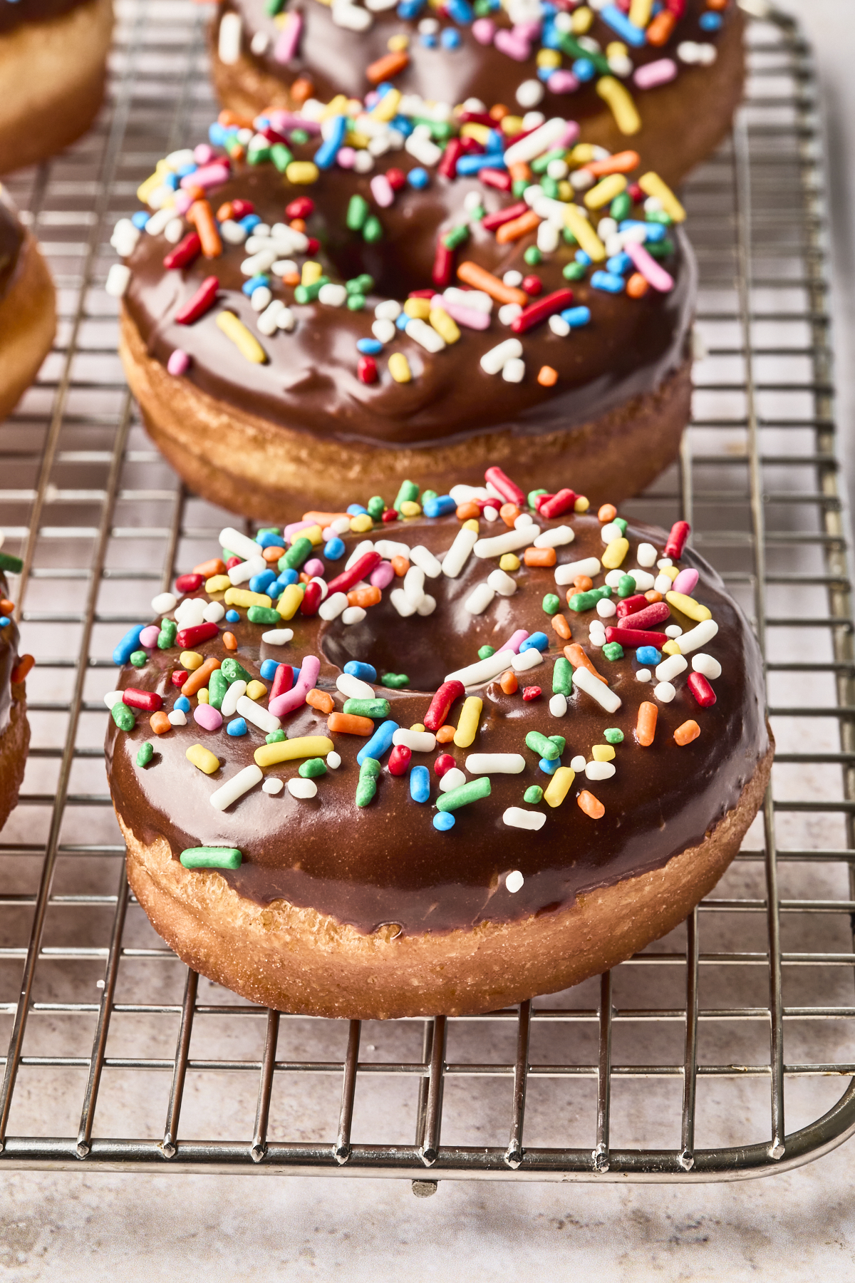 A chocolate-glazed donut topped with colorful rainbow sprinkles sits on a metal cooling rack, with other similar donuts in the background.