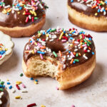 Chocolate-glazed donuts topped with colorful sprinkles sit on a light surface. One donut in the foreground has a bite taken out of it, with sprinkles scattered around.