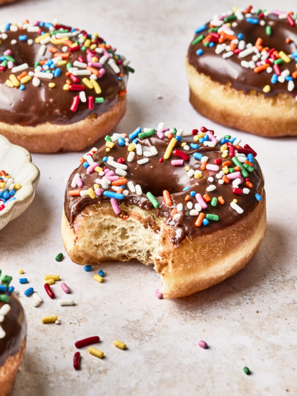 Chocolate-glazed donuts topped with colorful sprinkles sit on a light surface. One donut in the foreground has a bite taken out of it, with sprinkles scattered around.