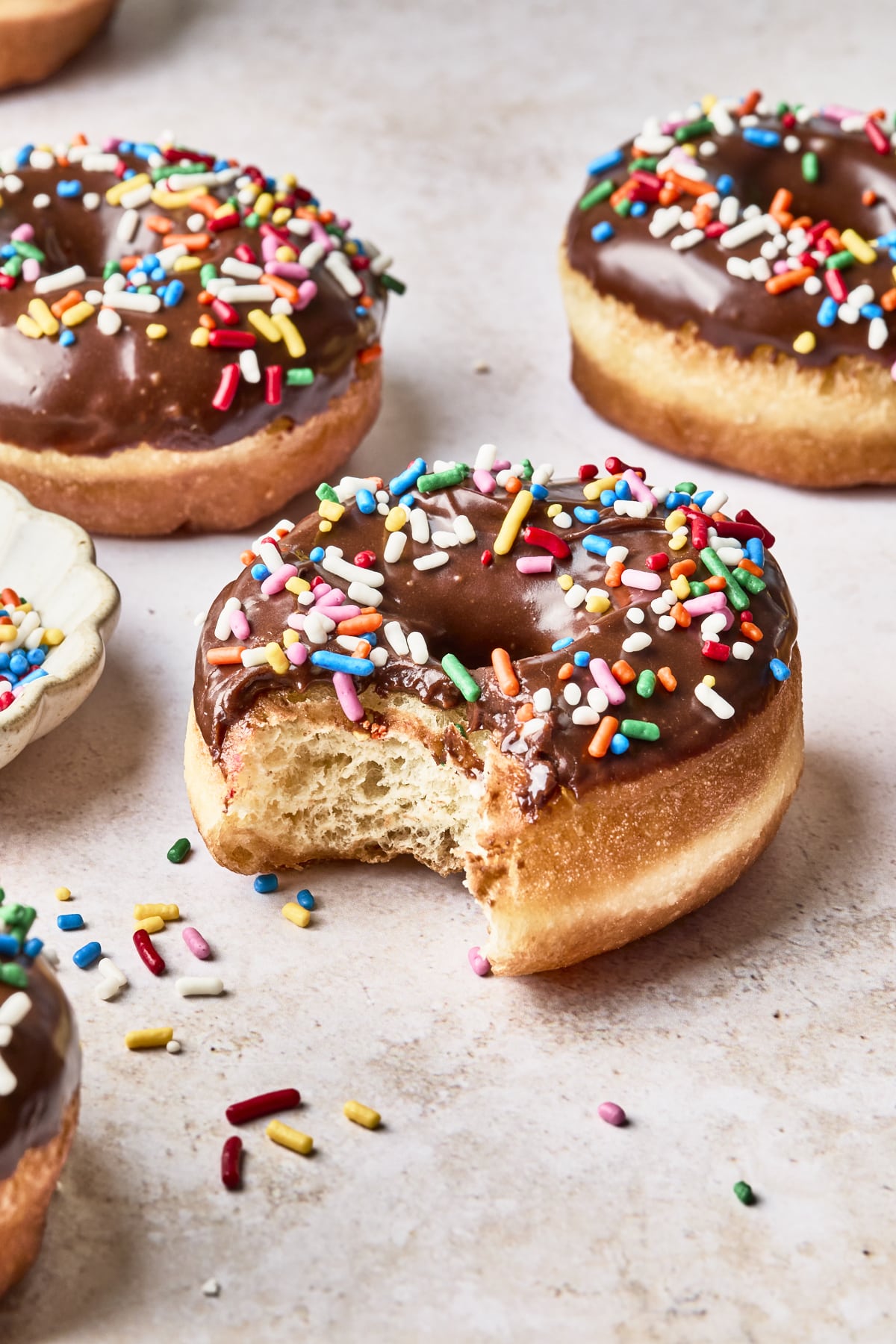 Chocolate-glazed donuts topped with colorful sprinkles sit on a light surface. One donut in the foreground has a bite taken out of it, with sprinkles scattered around.
