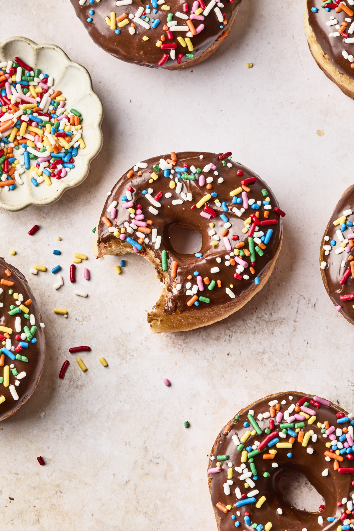 Chocolate-glazed donuts with colorful sprinkles are arranged on a light surface. One donut has a bite taken out. A small bowl of rainbow sprinkles sits nearby, with some sprinkles scattered around.