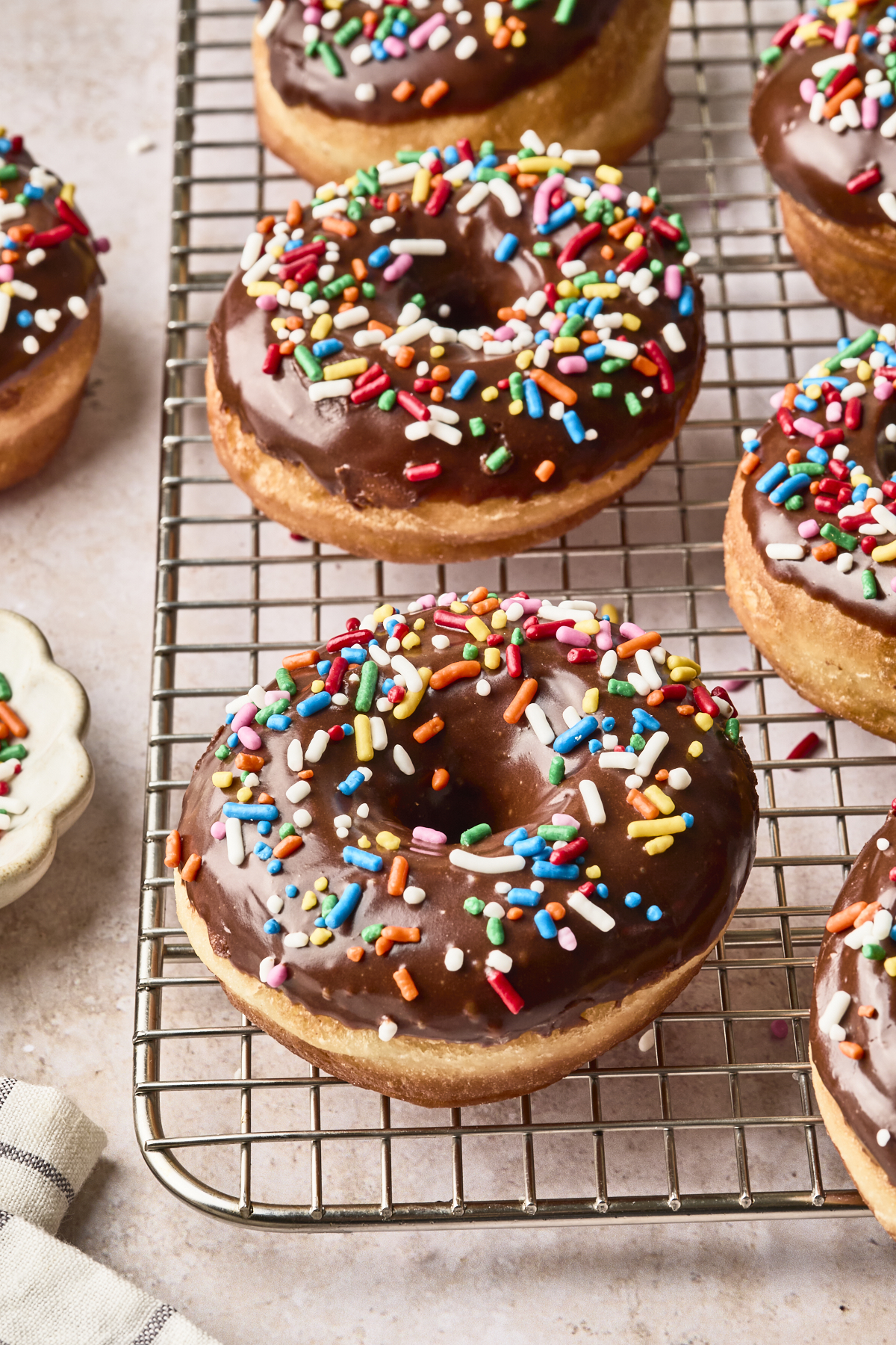 Chocolate-glazed donuts topped with colorful sprinkles are cooling on a wire rack. The donuts have a golden-brown color, and the rack sits on a light-colored countertop.