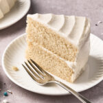 A slice of two-layer vanilla cake with white frosting sits on a white plate next to a silver fork. Colorful sprinkles are scattered on the light purple surface nearby.