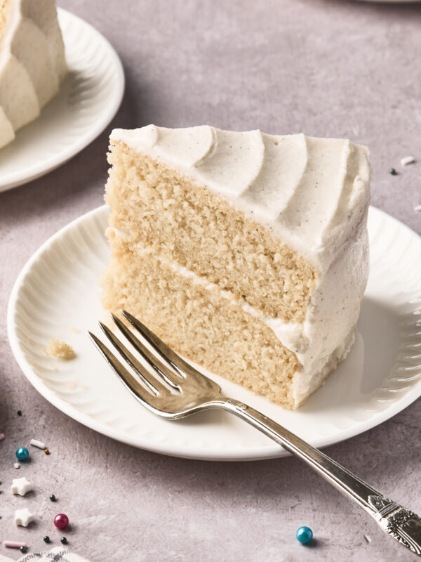 A slice of two-layer vanilla cake with white frosting sits on a white plate next to a silver fork. Colorful sprinkles are scattered on the light purple surface nearby.