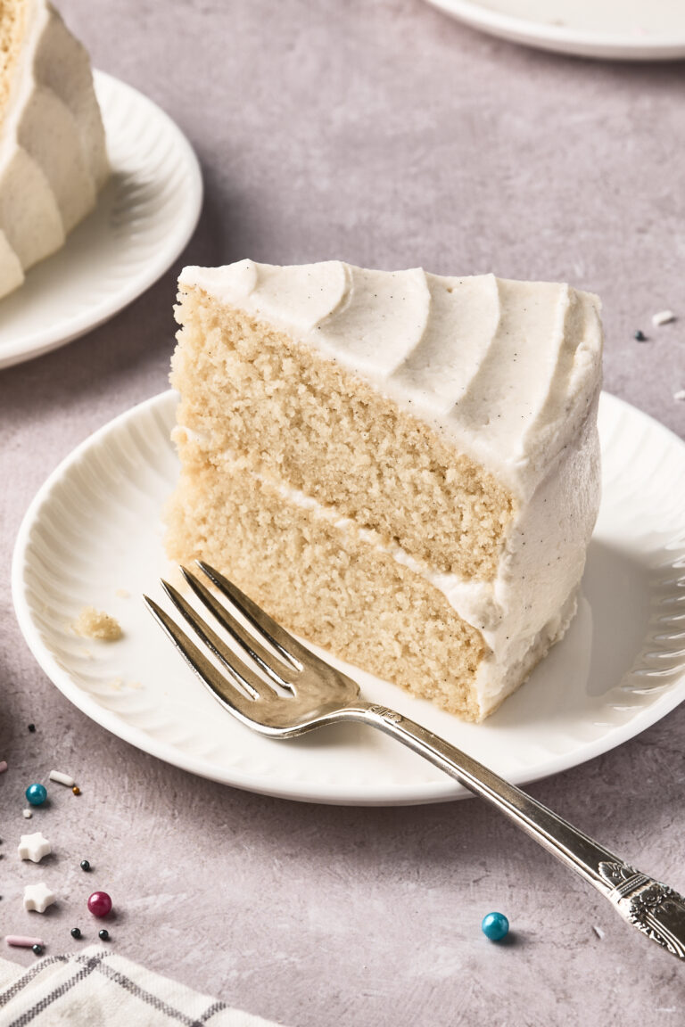 A slice of two-layer vanilla cake with white frosting sits on a white plate next to a silver fork. Colorful sprinkles are scattered on the light purple surface nearby.