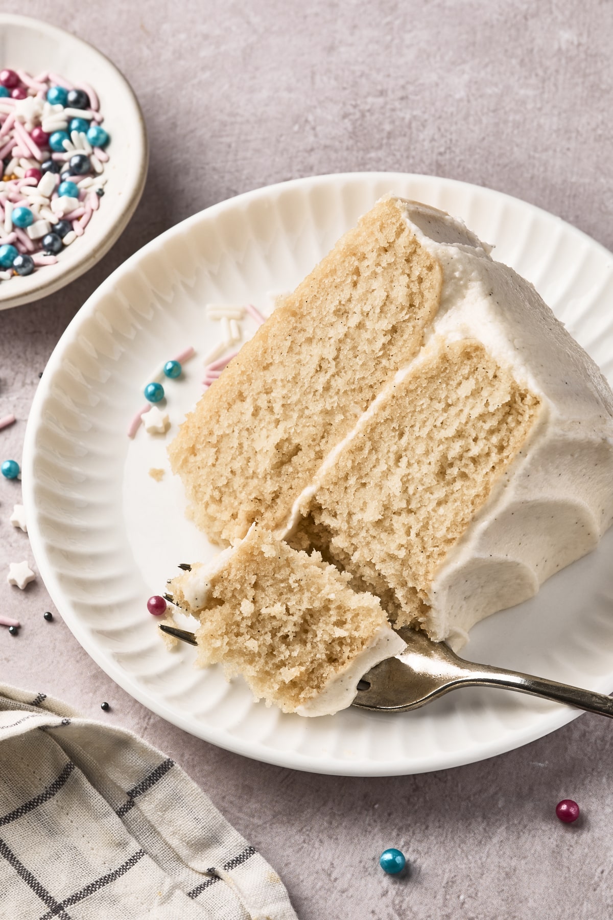 A slice of vanilla layer cake with creamy frosting on a white plate. A fork holds a bite of cake, and colorful sprinkles are scattered nearby, with a bowl of sprinkles and a striped cloth in the background.