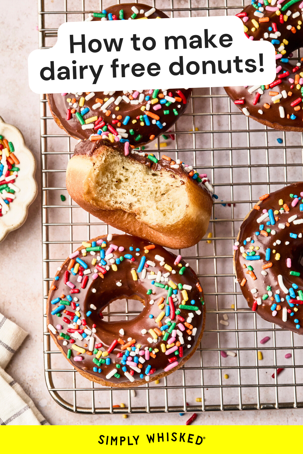 A cooling rack holds dairy free donuts with chocolate glaze and colorful sprinkles. One donut has a bite taken out. Text above reads, How to make dairy free donuts! with Simply Whisked at the bottom.