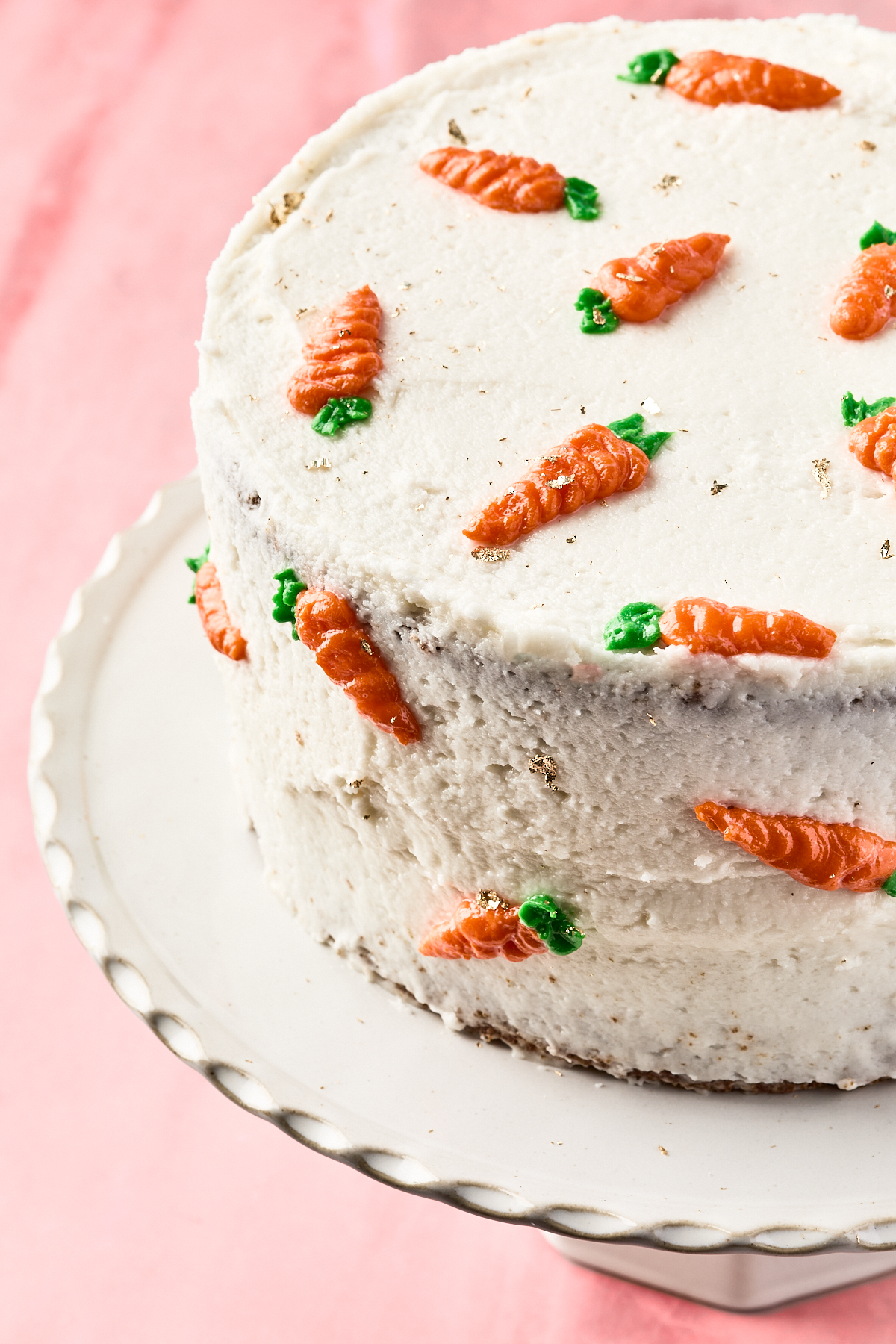 A round carrot cake covered in white frosting, decorated with small orange frosting carrots and green leaves, displayed on a white scalloped cake stand against a pink background.