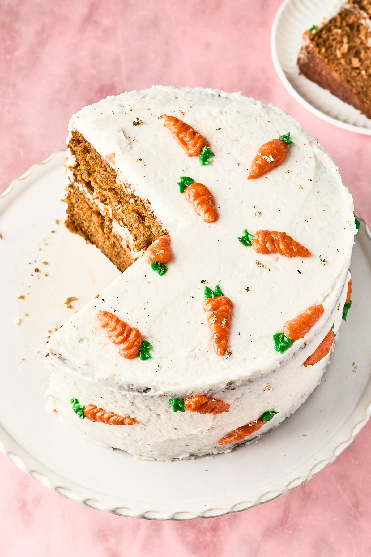 A round carrot cake with white frosting, decorated with small orange and green icing carrots, sits on a white cake stand. A large slice has been cut and slightly removed. The background is pink.