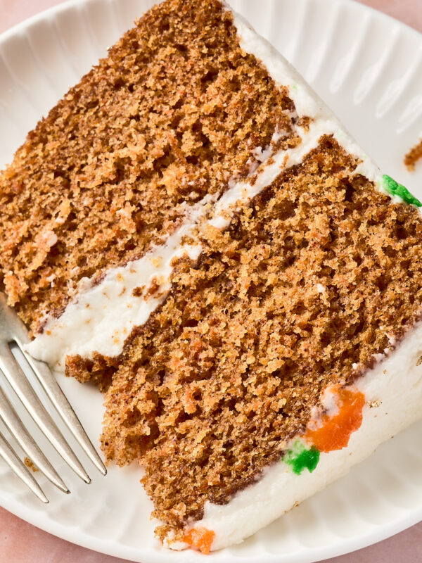 A close-up of two slices of carrot cake with cream cheese frosting on a white plate, accompanied by a silver fork. Small bits of orange and green frosting are visible on the cake edge.