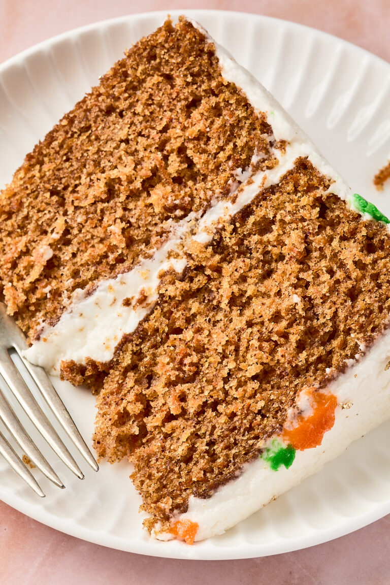 A close-up of two slices of carrot cake with cream cheese frosting on a white plate, accompanied by a silver fork. Small bits of orange and green frosting are visible on the cake edge.