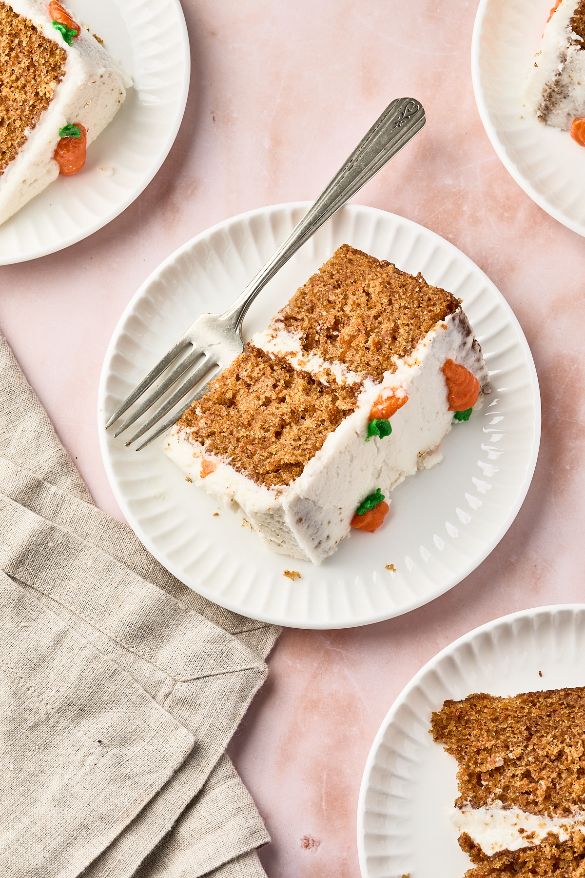 A plate with two slices of frosted carrot cake, garnished with small orange icing carrots, sits next to a fork on a pink surface with a beige napkin nearby. Other partial plates of cake are visible.