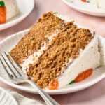 A close-up of a slice of layered carrot cake with white frosting and orange decorations on a white plate, with a fork beside it. Two more slices of cake are in the background on separate plates.