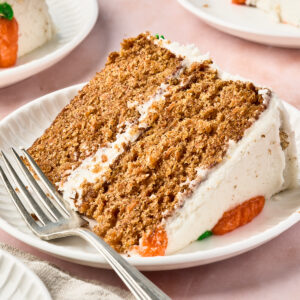 A close-up of a slice of layered carrot cake with white frosting and orange decorations on a white plate, with a fork beside it. Two more slices of cake are in the background on separate plates.