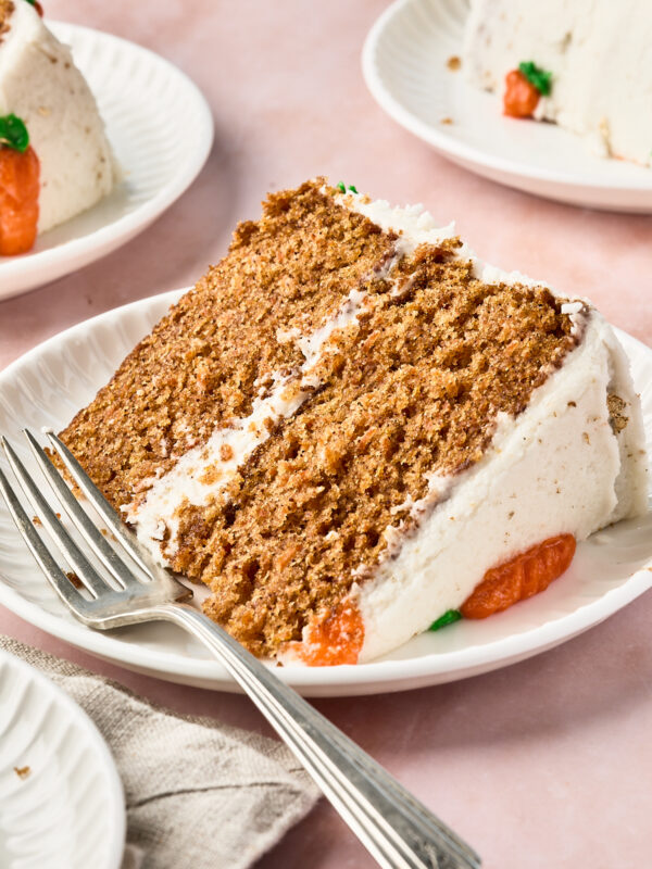 A close-up of a slice of layered carrot cake with white frosting and orange decorations on a white plate, with a fork beside it. Two more slices of cake are in the background on separate plates.