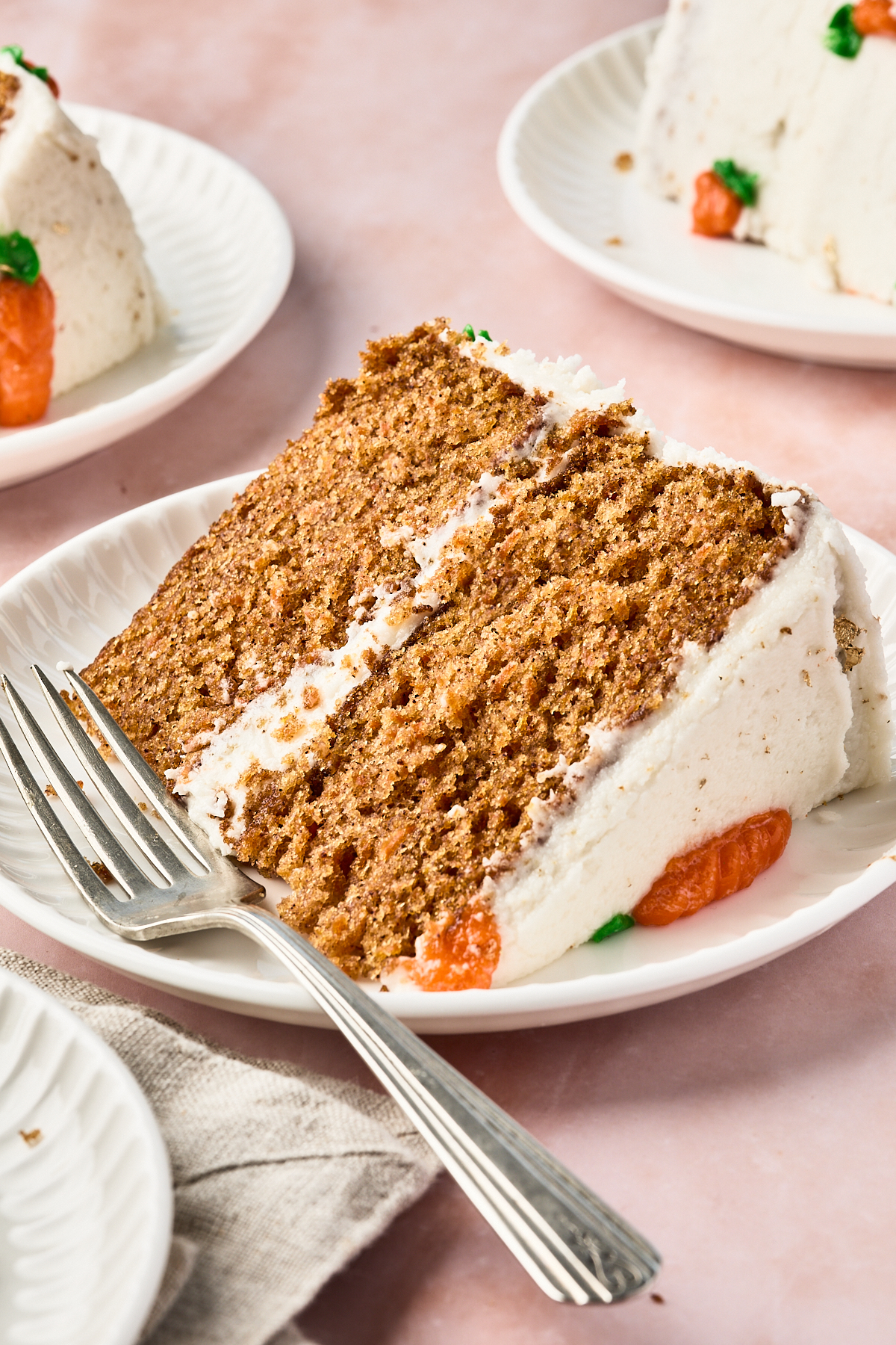 A close-up of a slice of layered carrot cake with white frosting and orange decorations on a white plate, with a fork beside it. Two more slices of cake are in the background on separate plates.